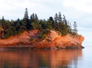 Photo of the cliffs at St Martins, New Brunswick.  Calm water in the foreground, orange rocks reflecting the setting sun, topped by evergreen trees and a light blue cloudy sky.